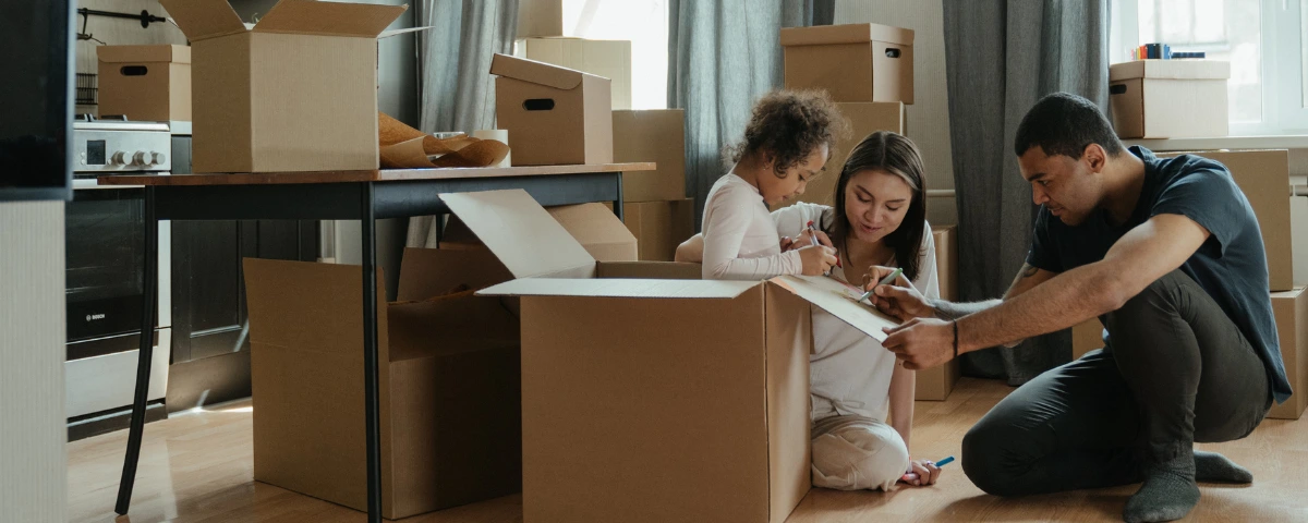 A diverse family sitting on the floor packing cardboard boxes and coloring, illustrating how to involve children in the moving process to reduce stress.
